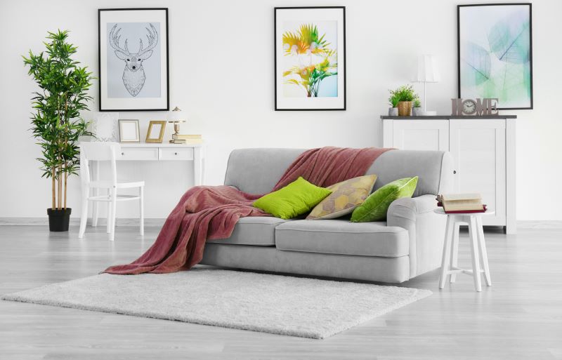 sofá no meio de sala de estar com piso vinílico de cimento queimado, decorada com tapete cinza, quadros, manta sobre o sofá, almofadas, plantas e escrivaninha